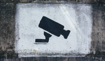 Mustvalge šabloonina maalitud turvakaamera ikoon karedal ja määrdunud betoonseinal.