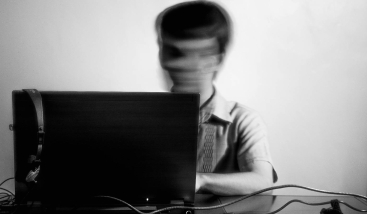 Mustvalge lähikaader sülearvuti taga istuvast inimesest, kelle nägu on tahtlikult hägustatud.